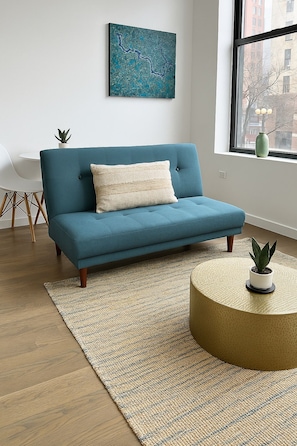 Un canapé de couleur turquoise avec un coussin texturé, un tapis tissé, une table basse ronde ornée d’une plante, et une vue sur le paysage urbain par la fenêtre.