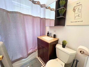 A bathroom with a shower curtain, a sink with a mirror above, a toilet, and a small shelf with plants. - Modern City Vibes | 1BR Stay (Chicago)