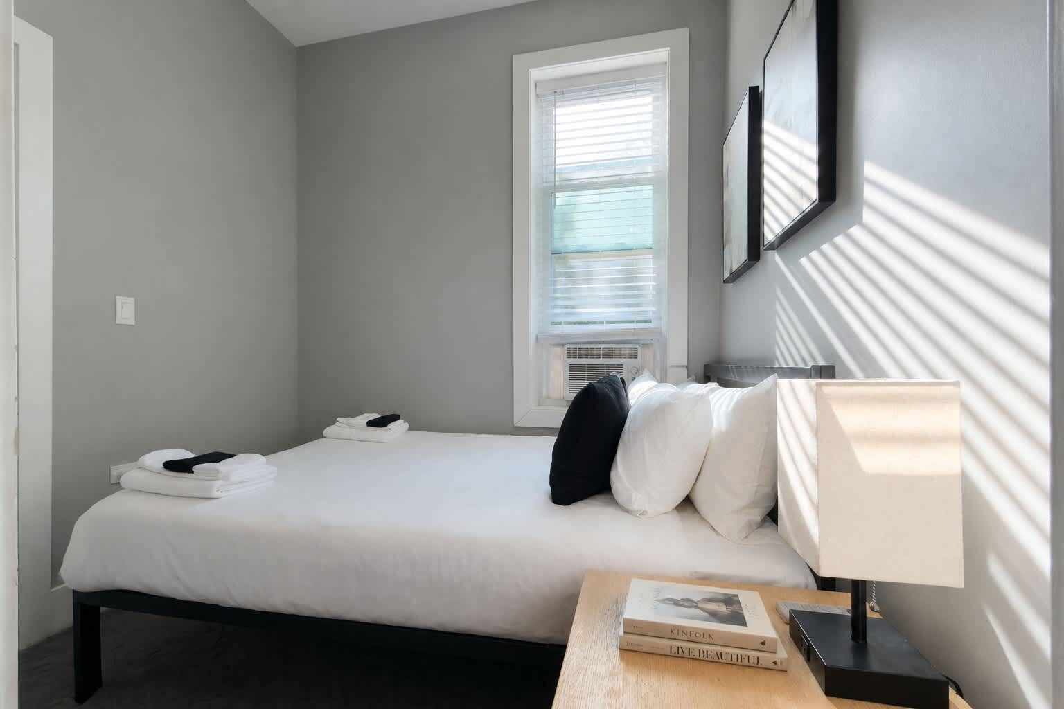 Une chambre d’hôtel comprenant un lit, une table de chevet avec une lampe, une fenêtre avec des stores et un cadre photo fixé au mur.