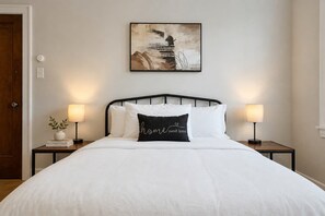Una cama bien hecha con ropa de cama blanca y un almohadón negro con el texto 'home sweet home', flanqueada por dos mesitas de noche con lámparas, en una habitación con una pared lisa y una puerta.