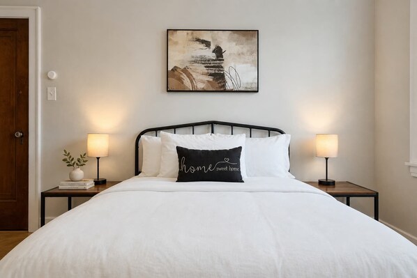Un letto rifatto con biancheria bianca e un cuscino nero con la scritta 'home sweet home', affiancato da due comodini con lampade, in una stanza con una parete spoglia e una porta.