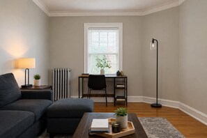 A living room with a sofa, a coffee table with books and a plant, a radiator, a desk with a chair, a floor lamp, and a window with blinds.