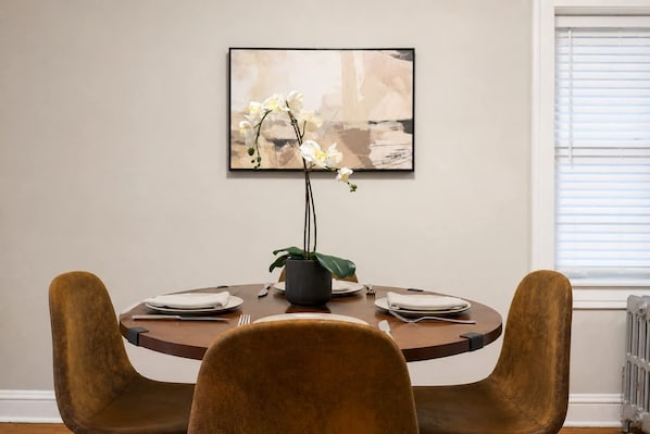 Un comedor con una mesa redonda, dos sillas, un florero con flores y una obra de arte enmarcada en la pared.