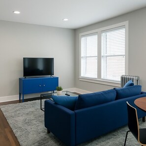 A modern living room with a blue sofa, a flat-screen TV on a blue cabinet, a small table with a plant, and a window with blinds. - 2BR Stylishly Apt in Lakeview (Chicago)