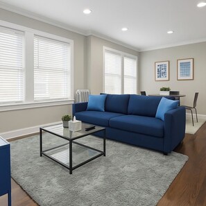 A living room with a blue sofa, a glass coffee table, and a grey rug. There are two windows with blinds, a radiator, and framed artwork on the wall. - 2BR Stylishly Apt in Lakeview (Chicago)