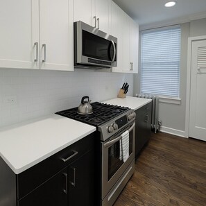 A kitchen with white cabinets, a stainless steel stove, a microwave, and a window with blinds. - 2BR Stylishly Apt in Lakeview (Chicago)