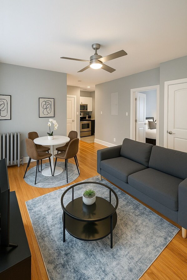 A modern living room with a grey sofa, a round coffee table, a dining table with chairs, a ceiling fan, and a kitchen in the background.