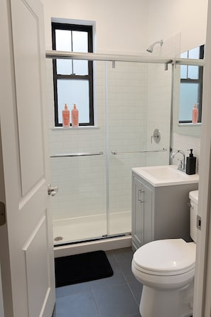 A modern bathroom with a glass shower enclosure, a toilet, a sink with a mirror, and a window with toiletries.