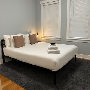 A neatly made bed with pillows and a stack of folded towels, a bedside lamp, and a window with blinds.