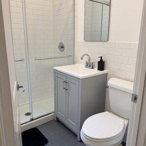Ein Badezimmer mit Duschkabine aus Glas, einem weißen Waschbecken mit Seifenspender und einer Toilette.