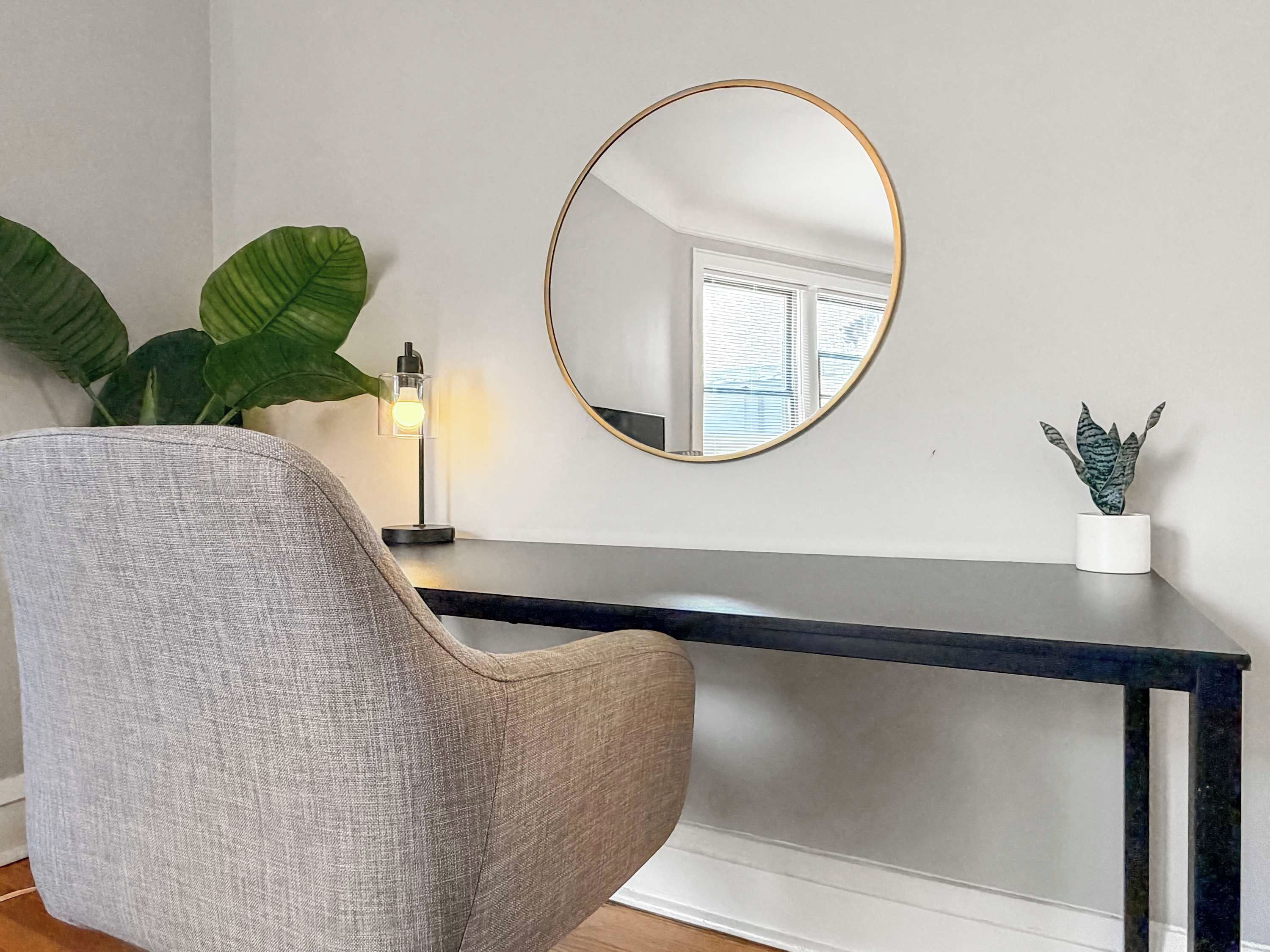 A room with a round mirror, a desk, a chair, a potted plant, and a small table with a plant.