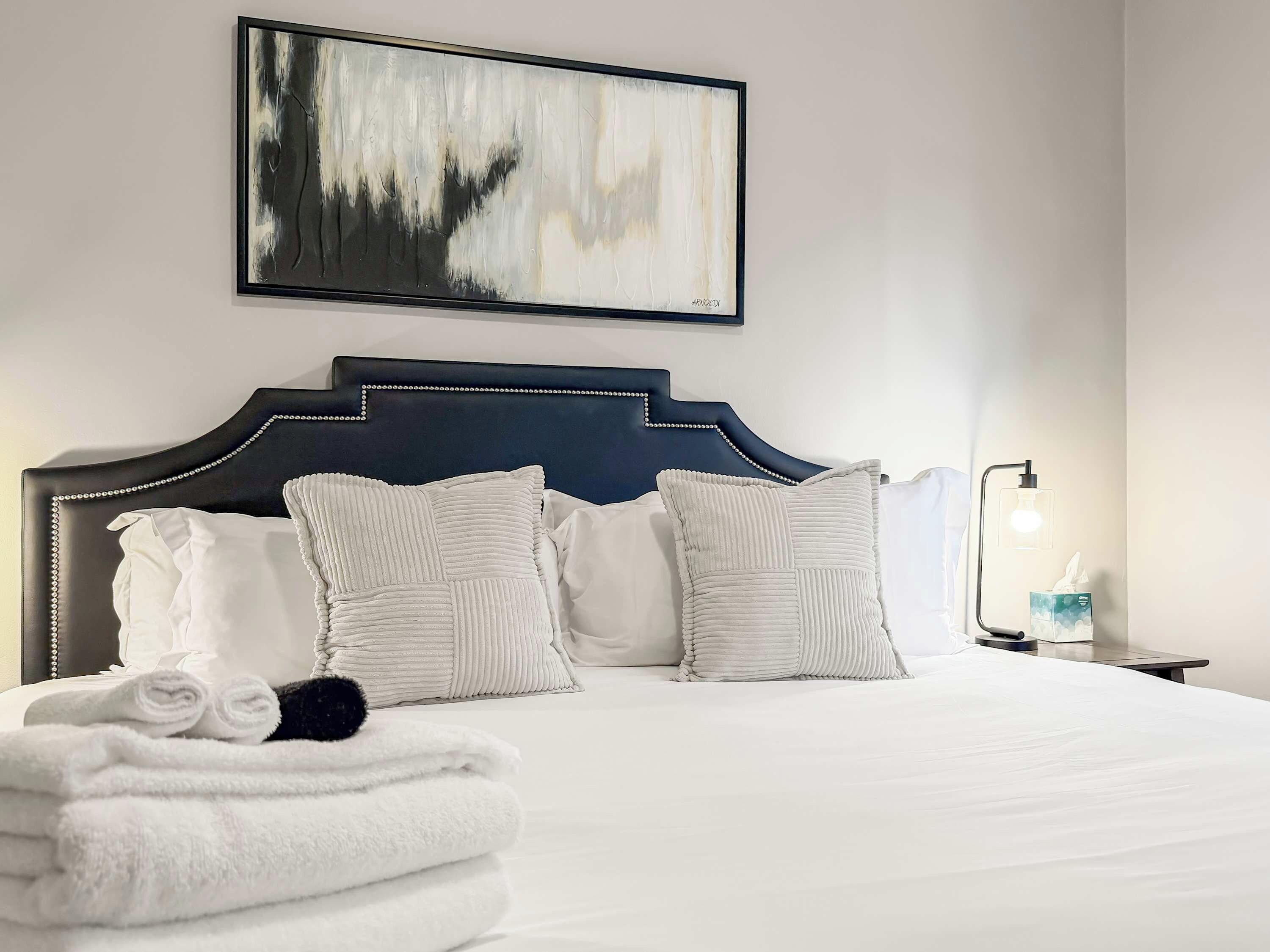 Cama bem arrumada com lençóis e almofadas brancas, cabeceira com detalhes em contas decorativas, pintura abstrata na parede e mesa de cabeceira com abajur e lenços.