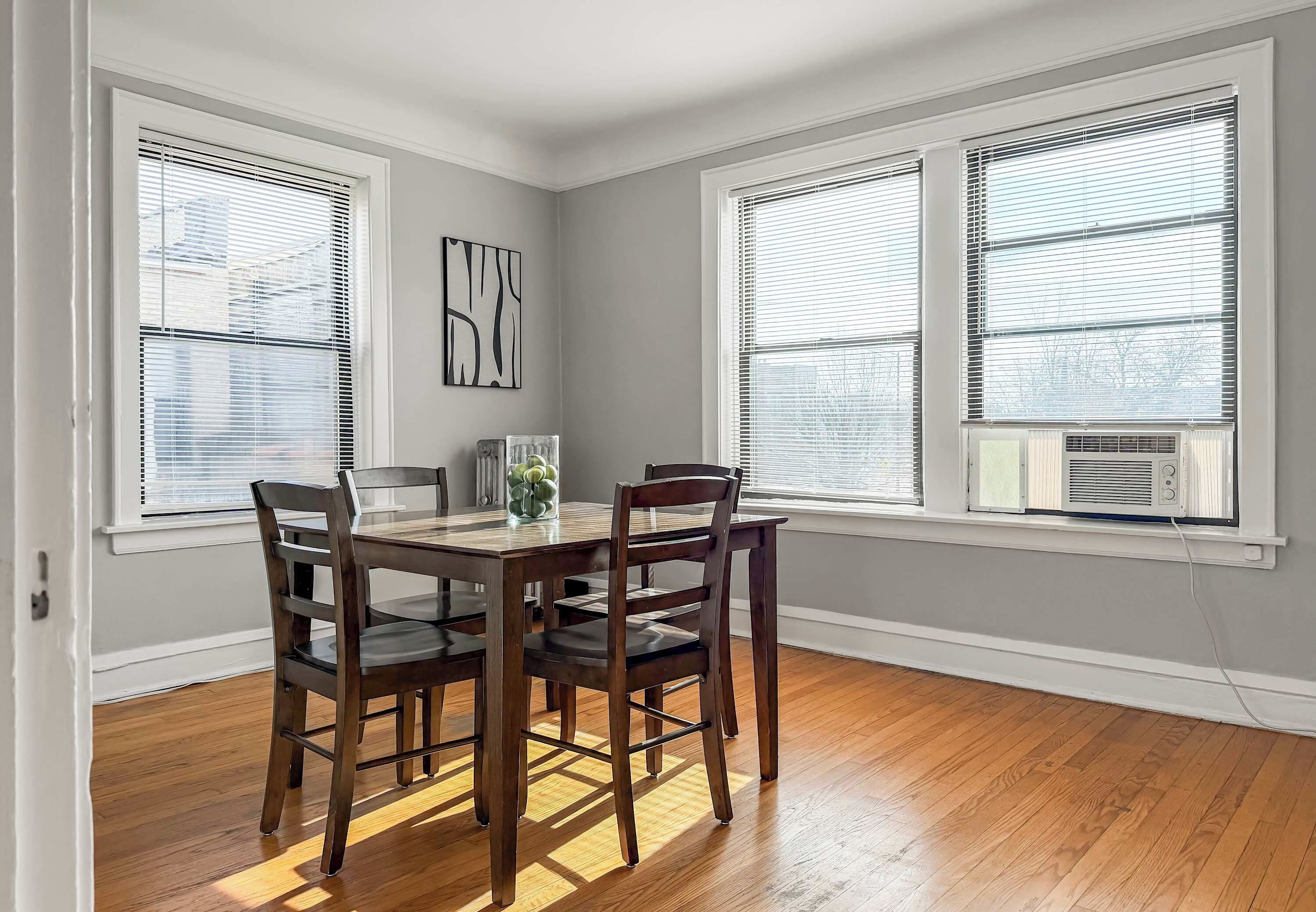 A dining area with a wooden table and chairs, large windows with blinds, and a radiator.