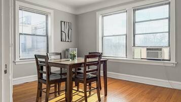 A dining area with a wooden table and chairs, large windows with blinds, and a radiator.