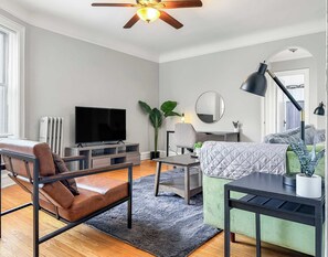 Un salon moderne avec un canapé en cuir, un fauteuil vert, un téléviseur, un ventilateur de plafond et une grande fenêtre.