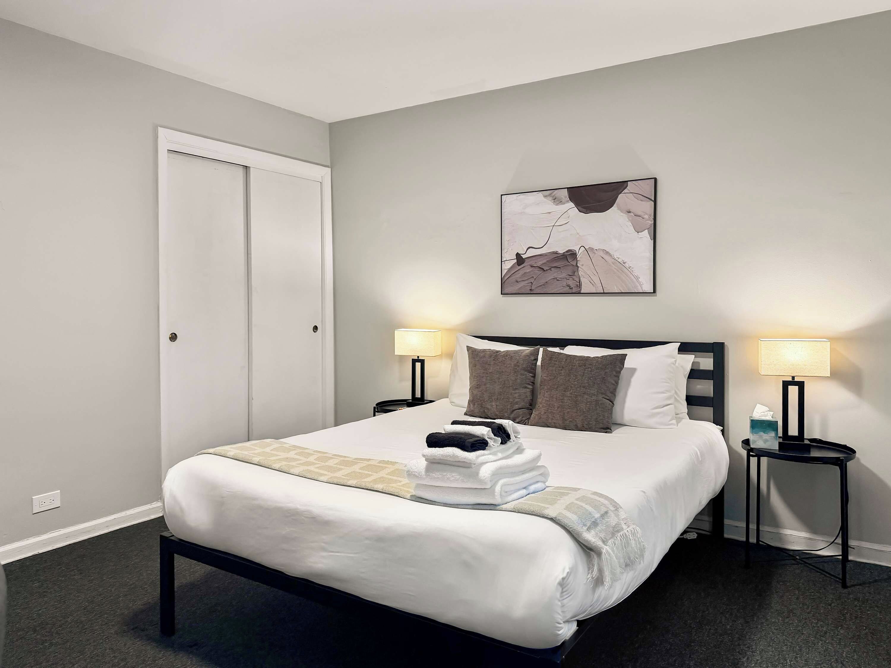 A neatly made bed with white linens and a folded towel, two bedside tables with lamps, a painting on the wall, and a closed door.