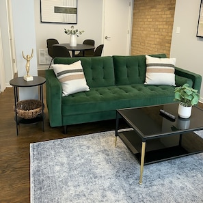A green sofa with two pillows, a small round table with a plant, a black coffee table, and a woven basket. - 2BR Calm & Aesthetic Apt (Chicago)