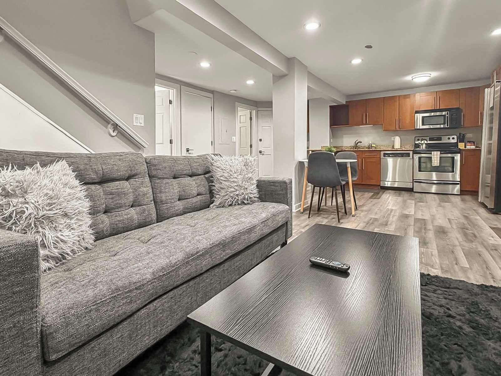 A modern living room with a grey sofa, a coffee table, and a kitchen area with wooden cabinets and stainless steel appliances.