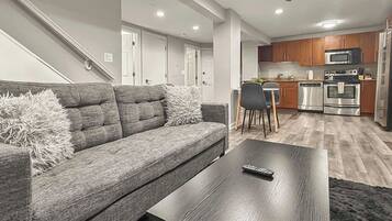 A modern living room with a grey sofa, a coffee table, and a kitchen area with wooden cabinets and stainless steel appliances.