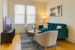 A living room with a teal sofa, a round coffee table, a TV on a stand, a lamp, and a painting on the wall.