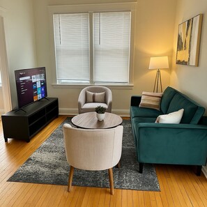 Ein Wohnzimmer mit Fernseher, einer Couch, einem Sessel, einer Lampe und einem Fenster mit Jalousien.