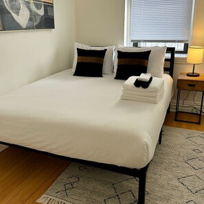 A neatly made bed with white bedding and two black and white pillows, a small bedside table with a lamp, and a patterned rug on a wooden floor.