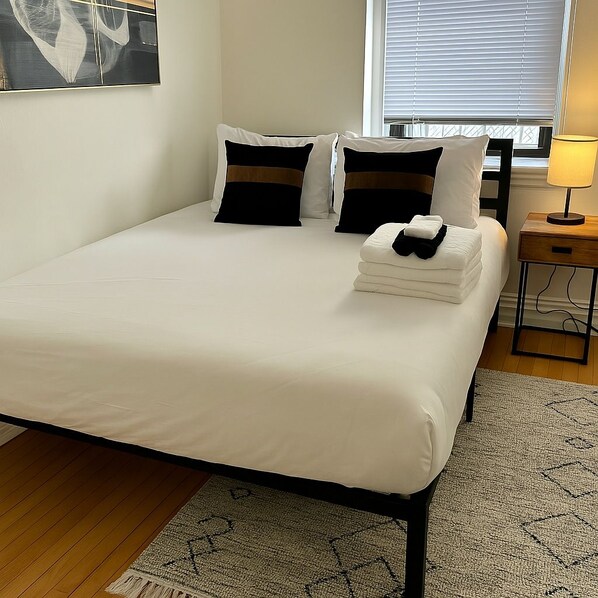 A neatly made bed with white bedding and two black and white pillows, a small bedside table with a lamp, and a patterned rug on a wooden floor.