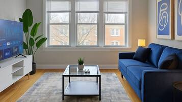 A living room with a blue sofa, a glass coffee table, a TV on a stand, and a large window with blinds.