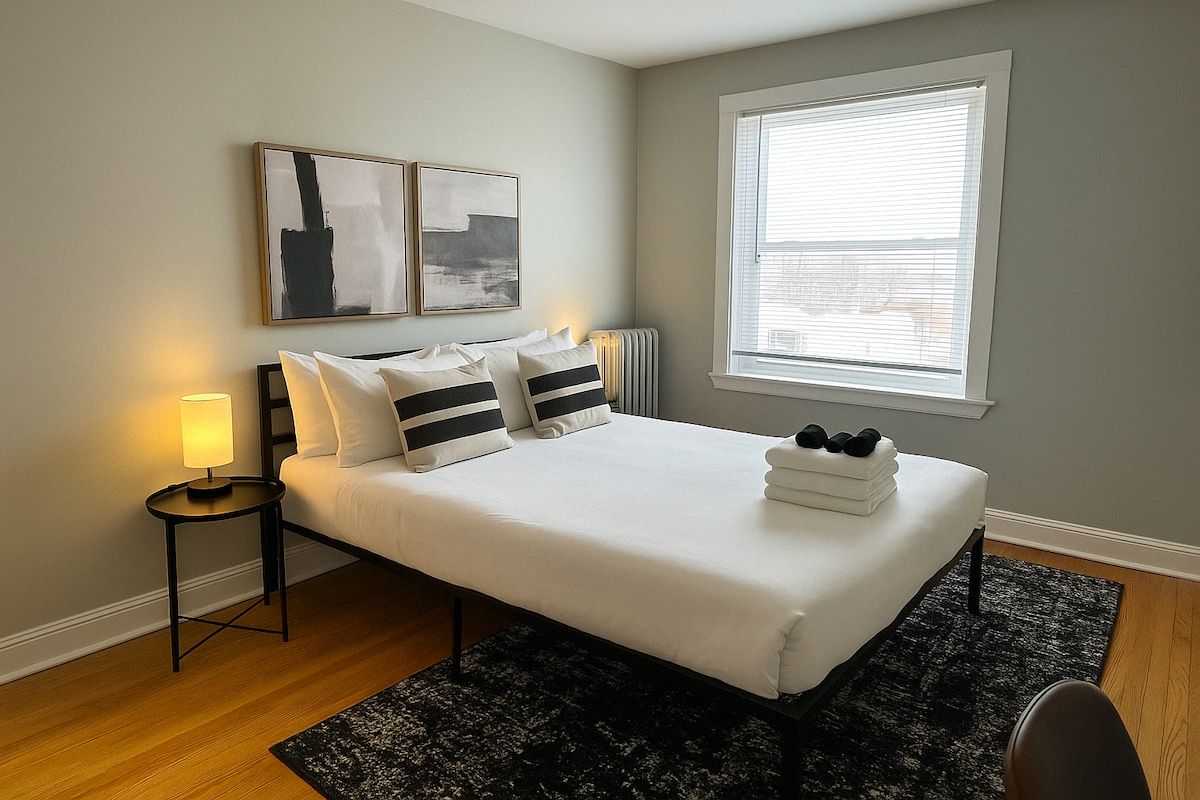 A bedroom with a bed, a bedside table with a lamp, a window with blinds, and artwork on the wall.