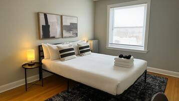 A bedroom with a bed, a bedside table with a lamp, a window with blinds, and artwork on the wall.