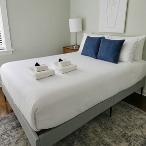 A neatly made bed with white linens and two folded towels, a bedside lamp, and a framed artwork on the wall.