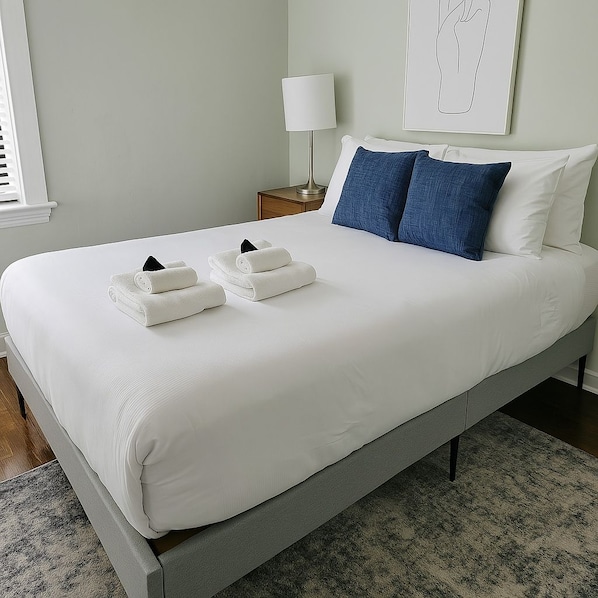 A neatly made bed with white linens and two folded towels, a bedside lamp, and a framed artwork on the wall.