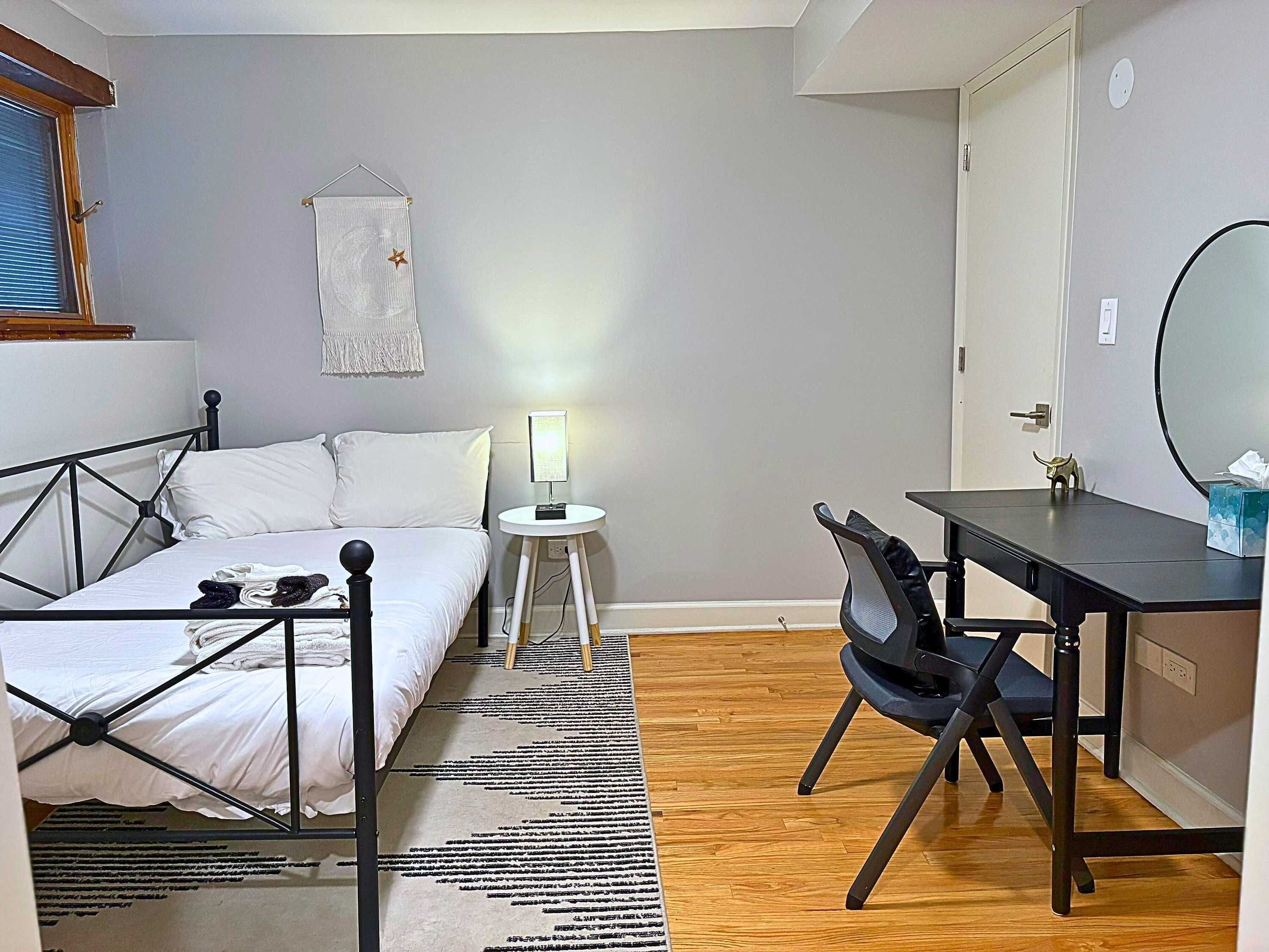 Un dormitorio con cama, mesita de noche, lámpara, silla, escritorio y espejo.