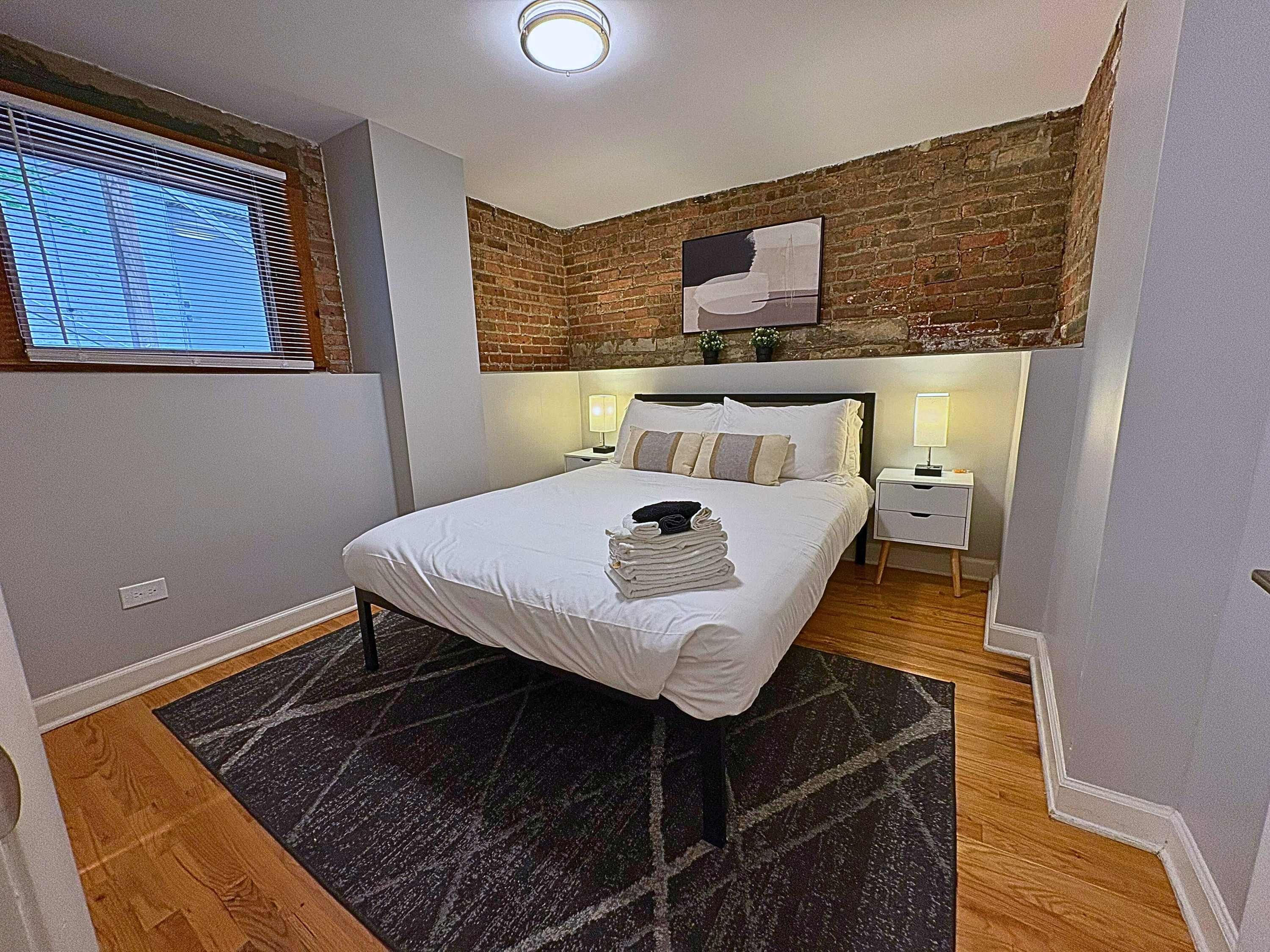 Un dormitorio moderno con pared de ladrillo decorativa, una cama grande con ropa de cama blanca, dos mesitas de noche con lámparas y una ventana con persianas.