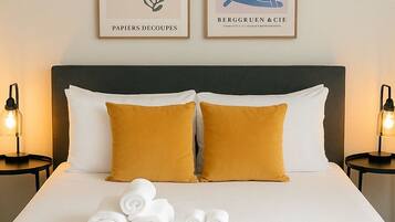 A neatly made bed with white and mustard pillows, two framed artworks above it, and rolled towels on the bed.