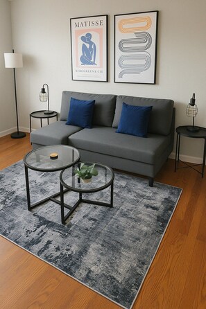 A living room with a grey sofa, two round glass tables, a grey patterned rug, and two framed artworks on the wall.