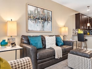 A modern living room with a leather sofa, a coffee table, and a patterned ottoman. There is a kitchen area with dark cabinets and a bar stool. - Luxury Living at the The Cays! Resort Pool/Spa, Steam, Gym. Waterfront Community! (Chandler)