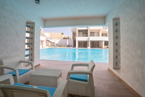 Et poolområde med fletningsmøbler og udsigt til en swimmingpool samt bygninger i baggrunden.