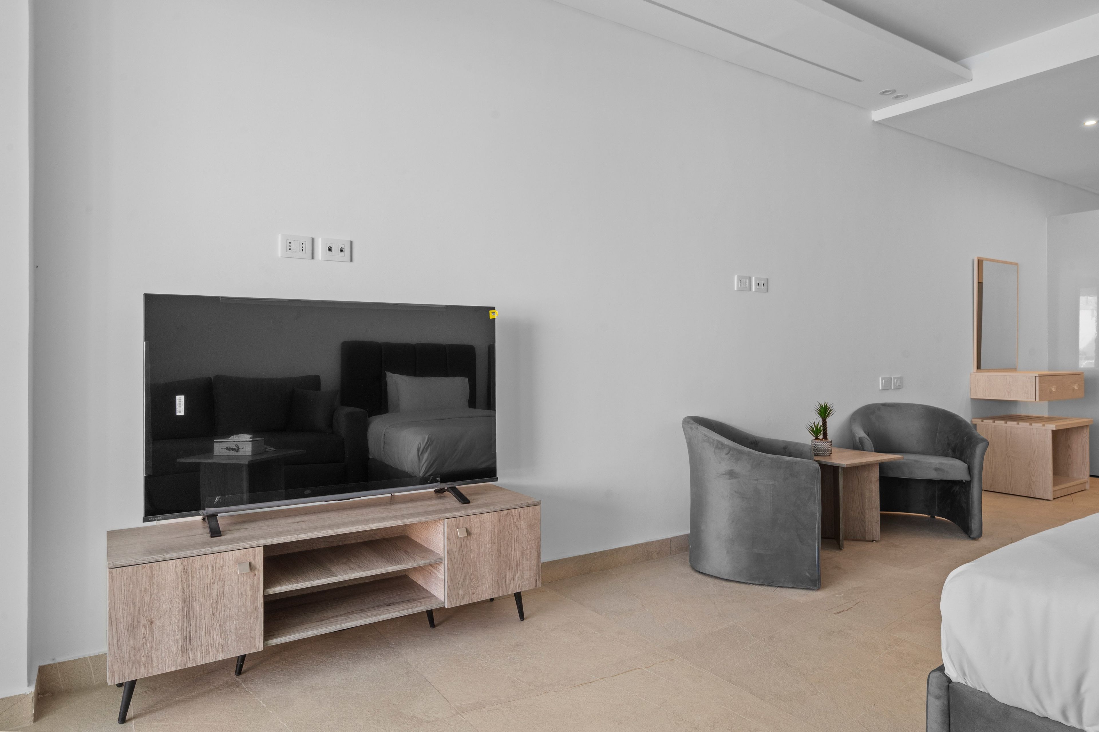 A modern hotel room with a large flat-screen TV, a wooden desk, a grey armchair, a small bedside table with a lamp, and a bed with a grey headboard.
