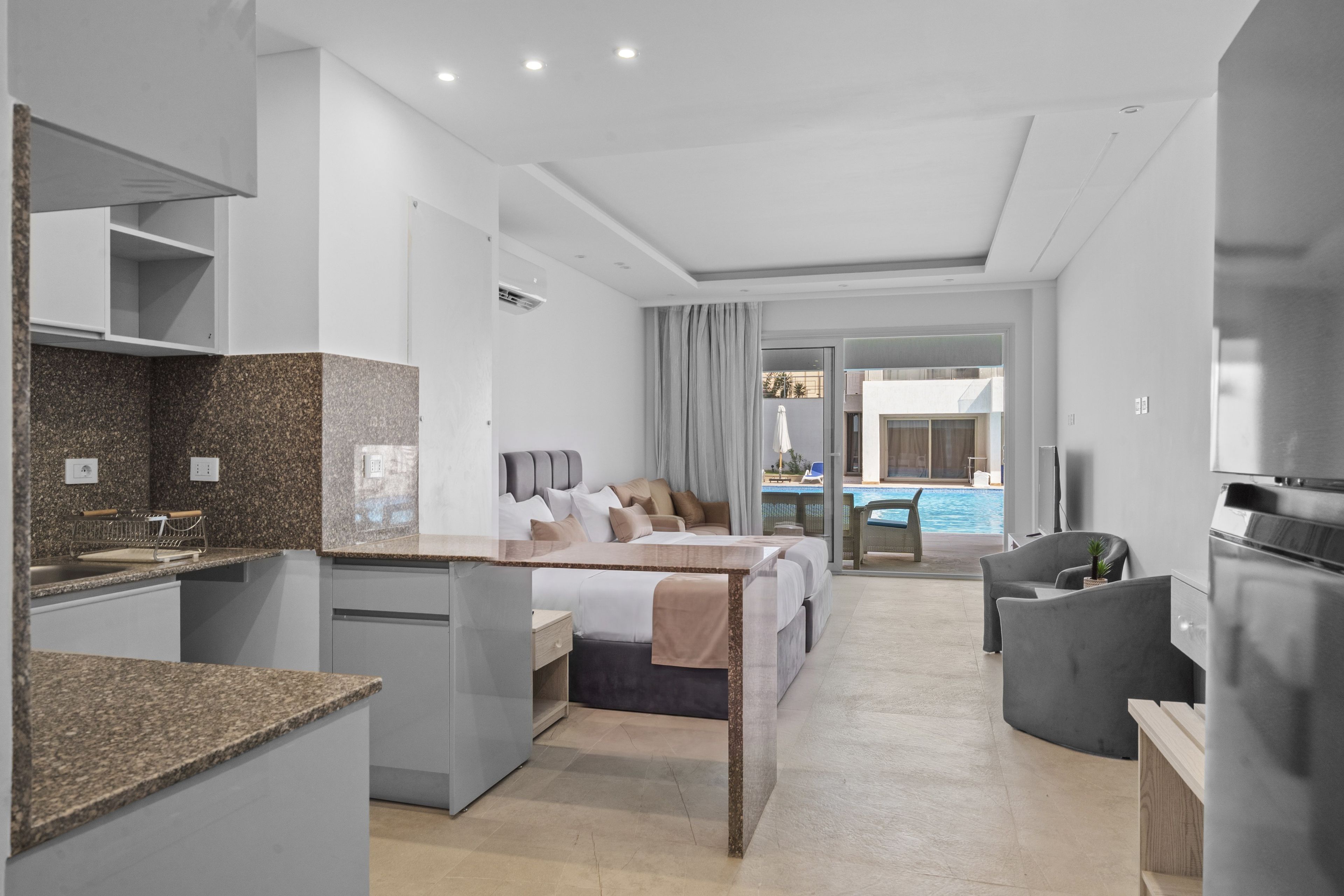 A modern hotel room with a kitchenette, a bed with a view of a pool, and a seating area.