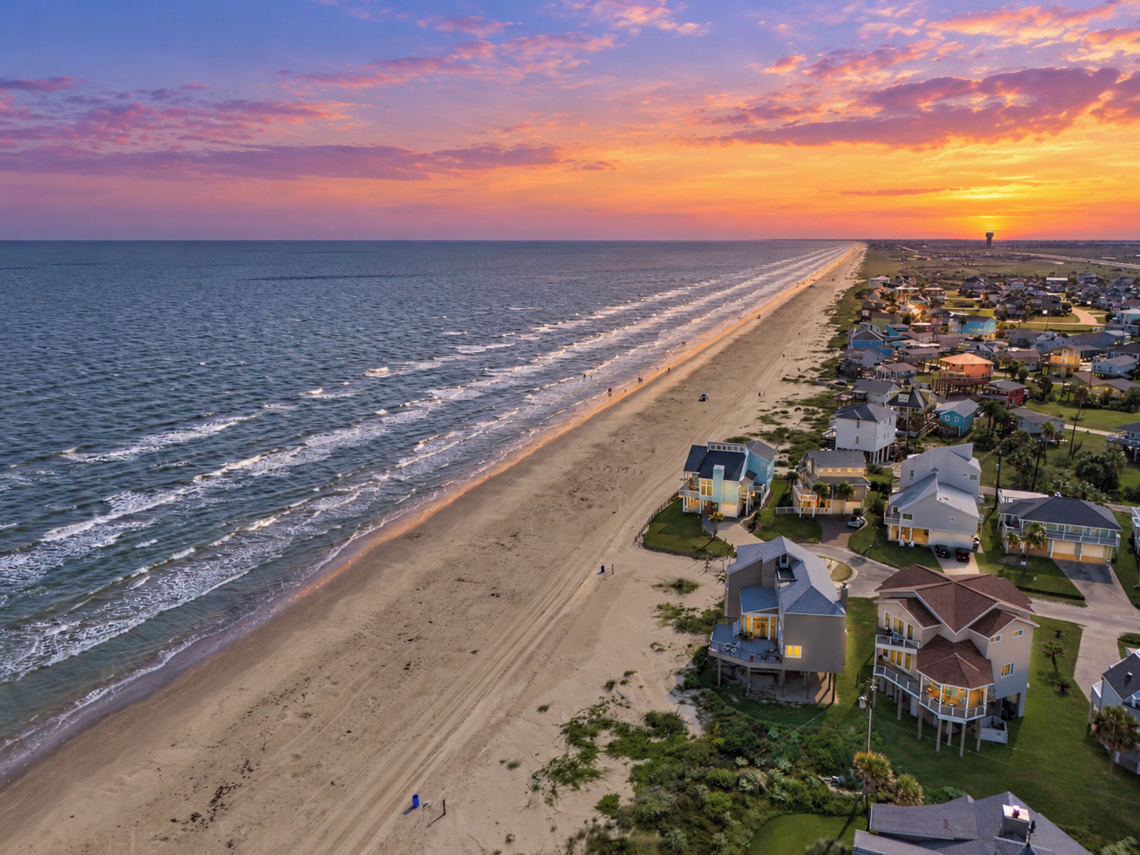 A coastal residential area with houses along the beach and a sunset in the background.
