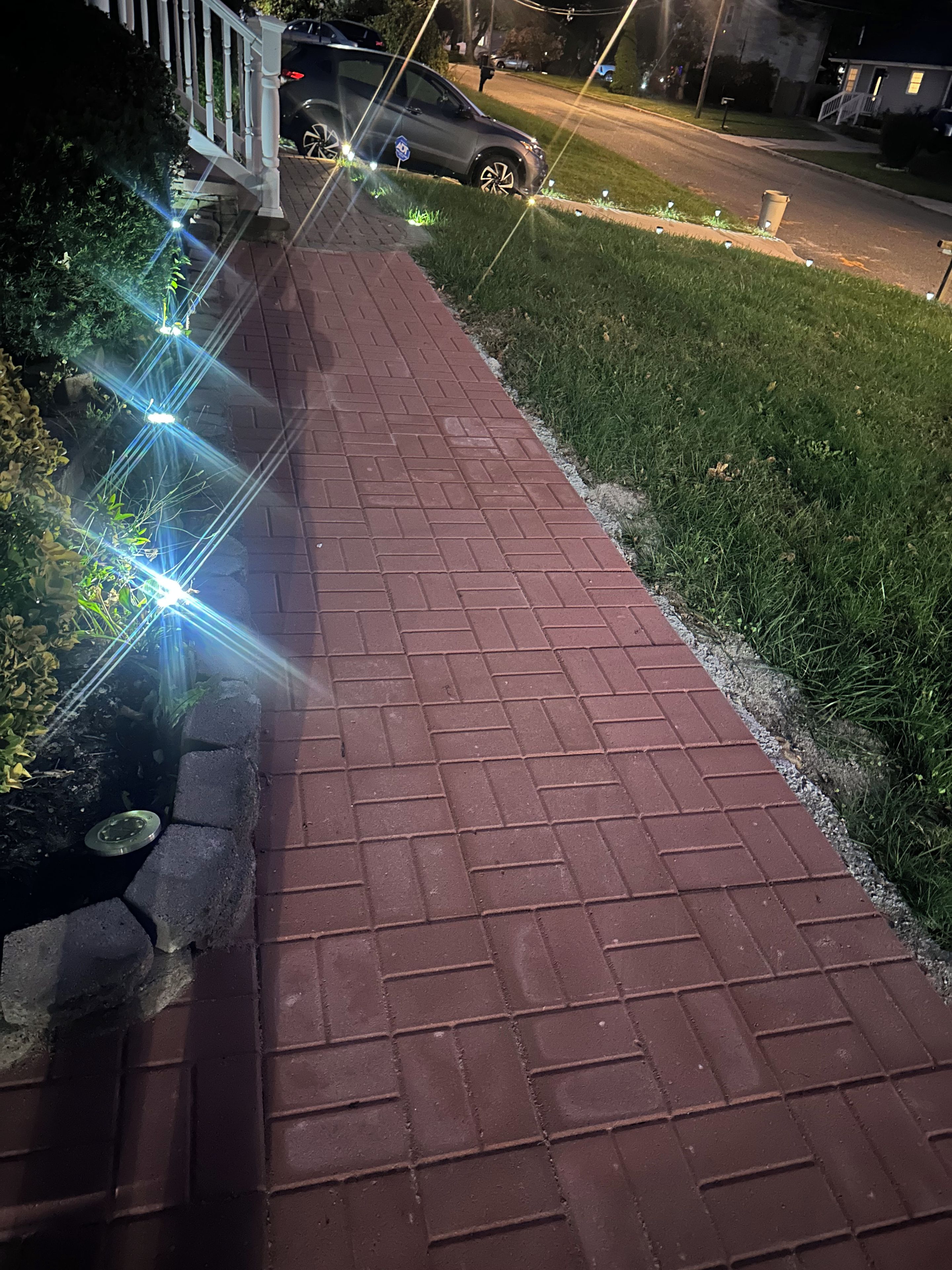 Un chemin pavé, dont le revêtement est constitué de briques disposées selon un motif, bordé de gazon et d’un trottoir.