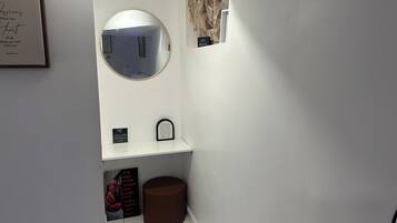 Un baño con tocador blanco, un espejo redondo, una cita inspiradora enmarcada, una ventana pequeña y un cubo de basura.