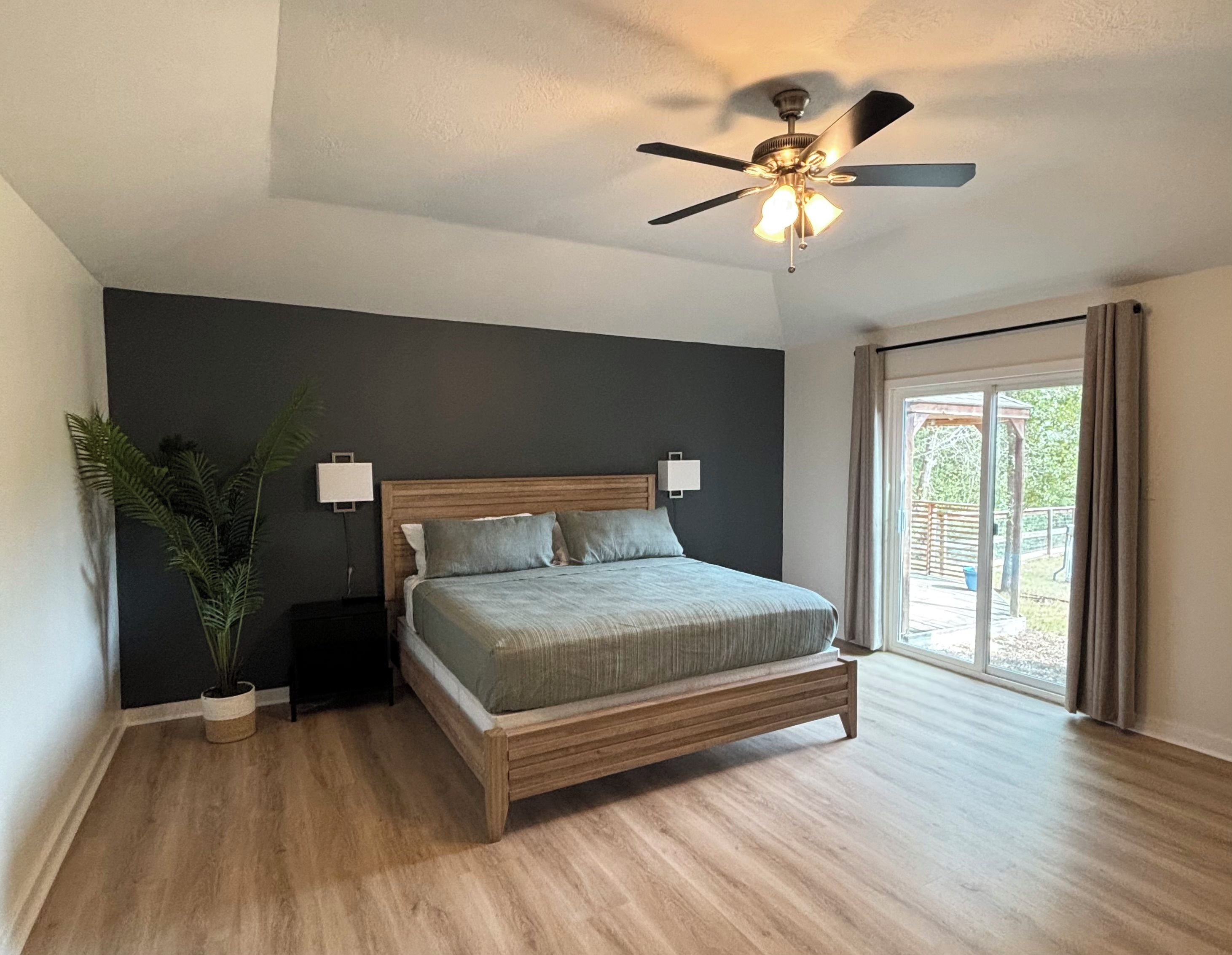 Une chambre à coucher avec un lit, un ventilateur de plafond, une table de chevet, une plante en pot et une fenêtre avec des rideaux.