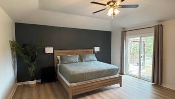 Une chambre à coucher avec un lit, un ventilateur de plafond, une table de chevet, une plante en pot et une fenêtre avec des rideaux.