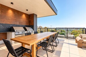 A modern outdoor kitchen with a grill, dining table, and chairs, overlooking a cityscape. - The Ember Retreat !! (San Diego)