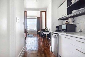 A narrow hallway leading to a kitchen with a dishwasher, a dining area with a table and chairs, and a view through a large window. - Heritage Hotel 1 Bedroom Apartment ! (Auckland CBD)