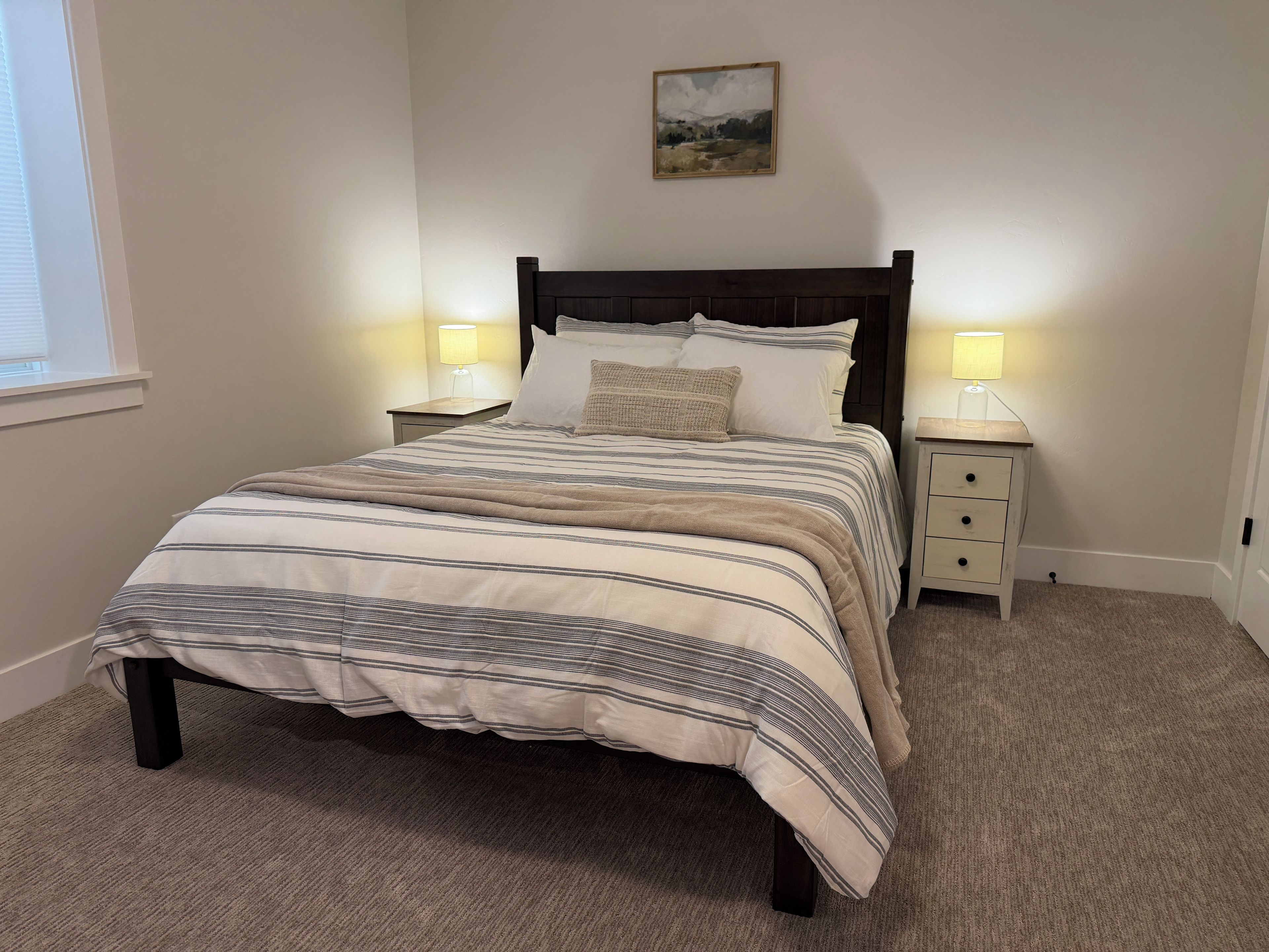 A bedroom with a bed, two bedside tables, a nightstand, a lamp, and a painting on the wall.
