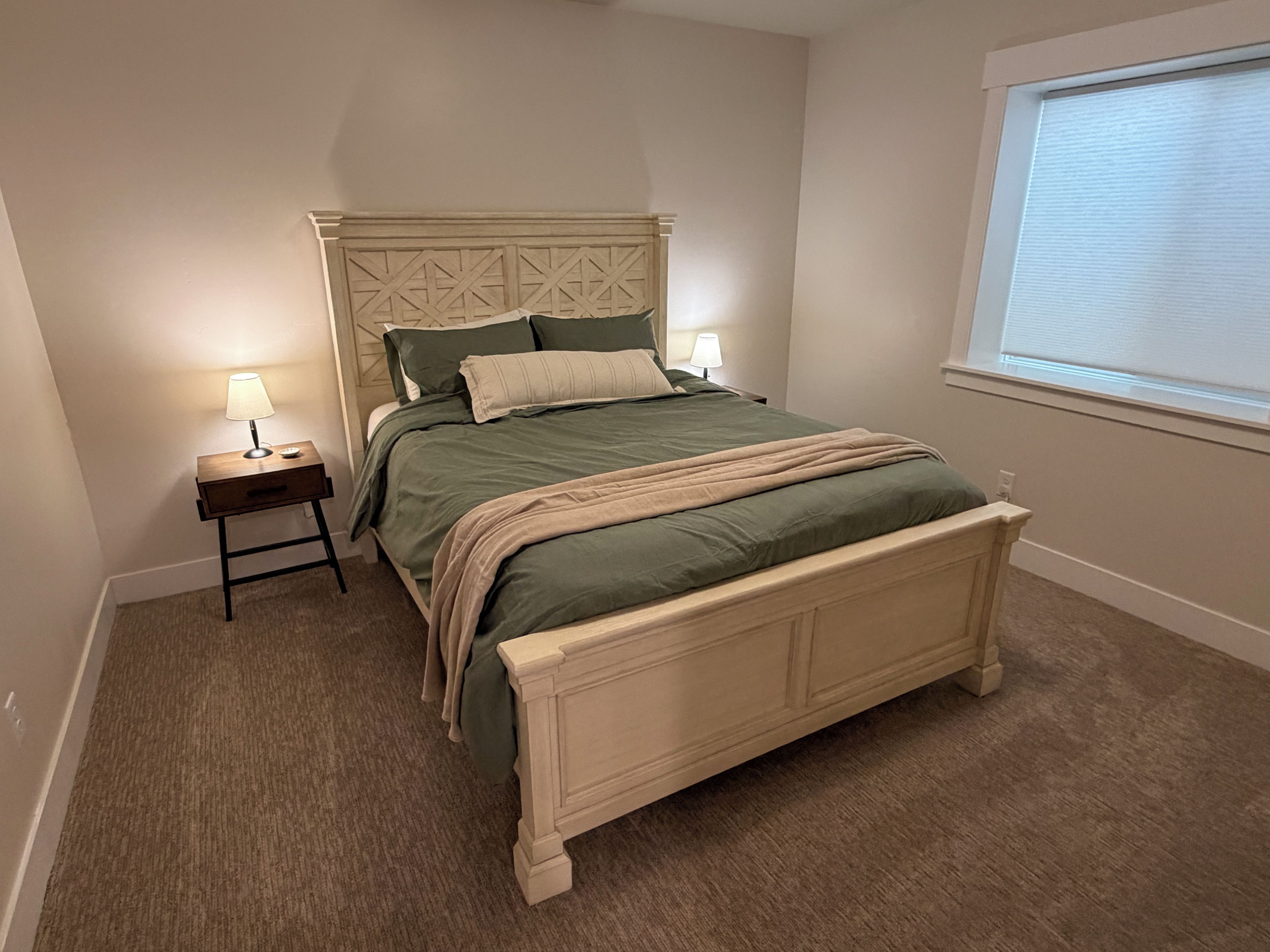 A bedroom with a bed, two bedside lamps, a window with blinds, and a carpeted floor.