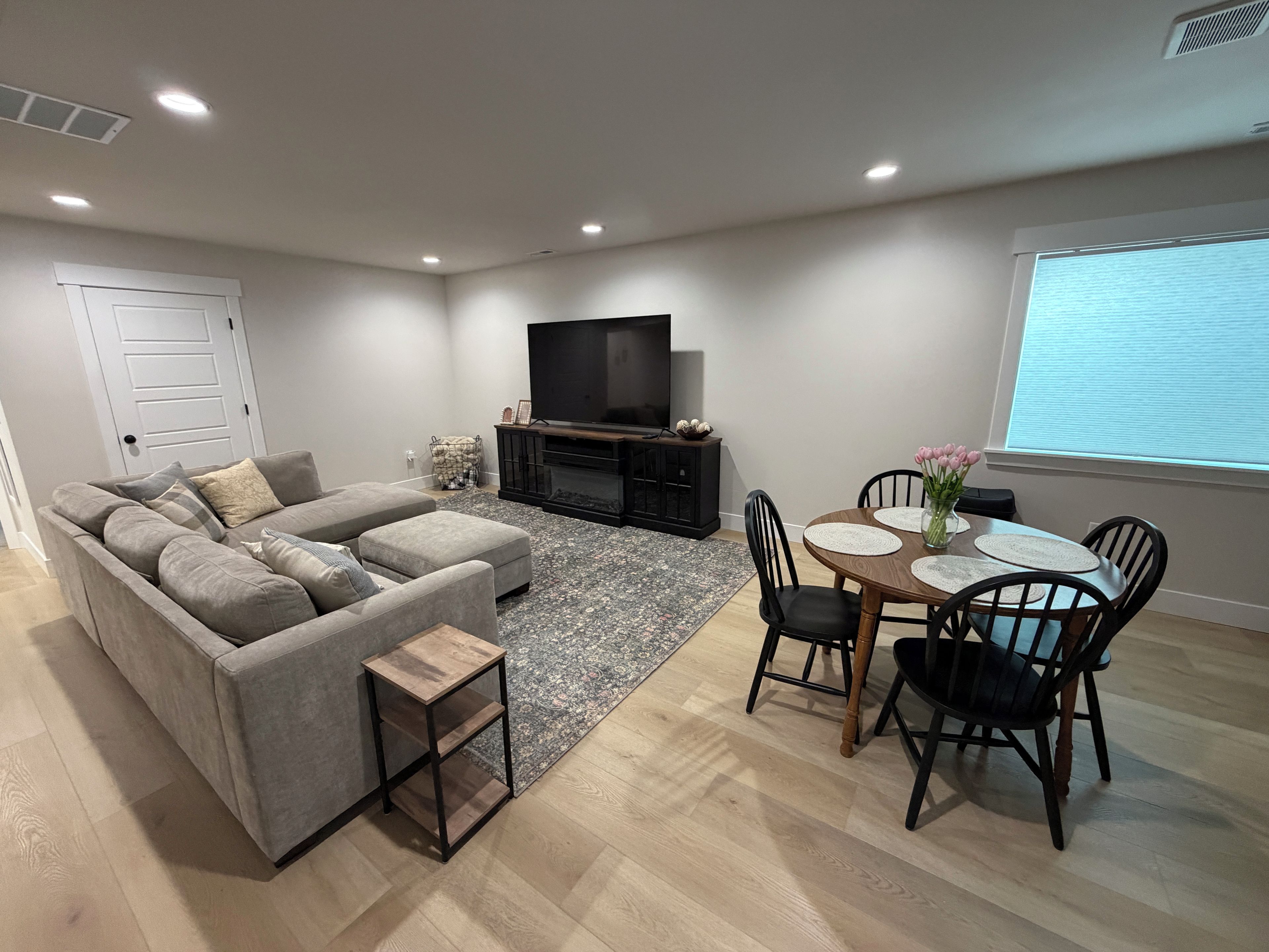 A modern living room with a grey sectional sofa, a small wooden side table, a dining area with a round table and chairs, a flat-screen TV on a black stand, and a window with blinds.